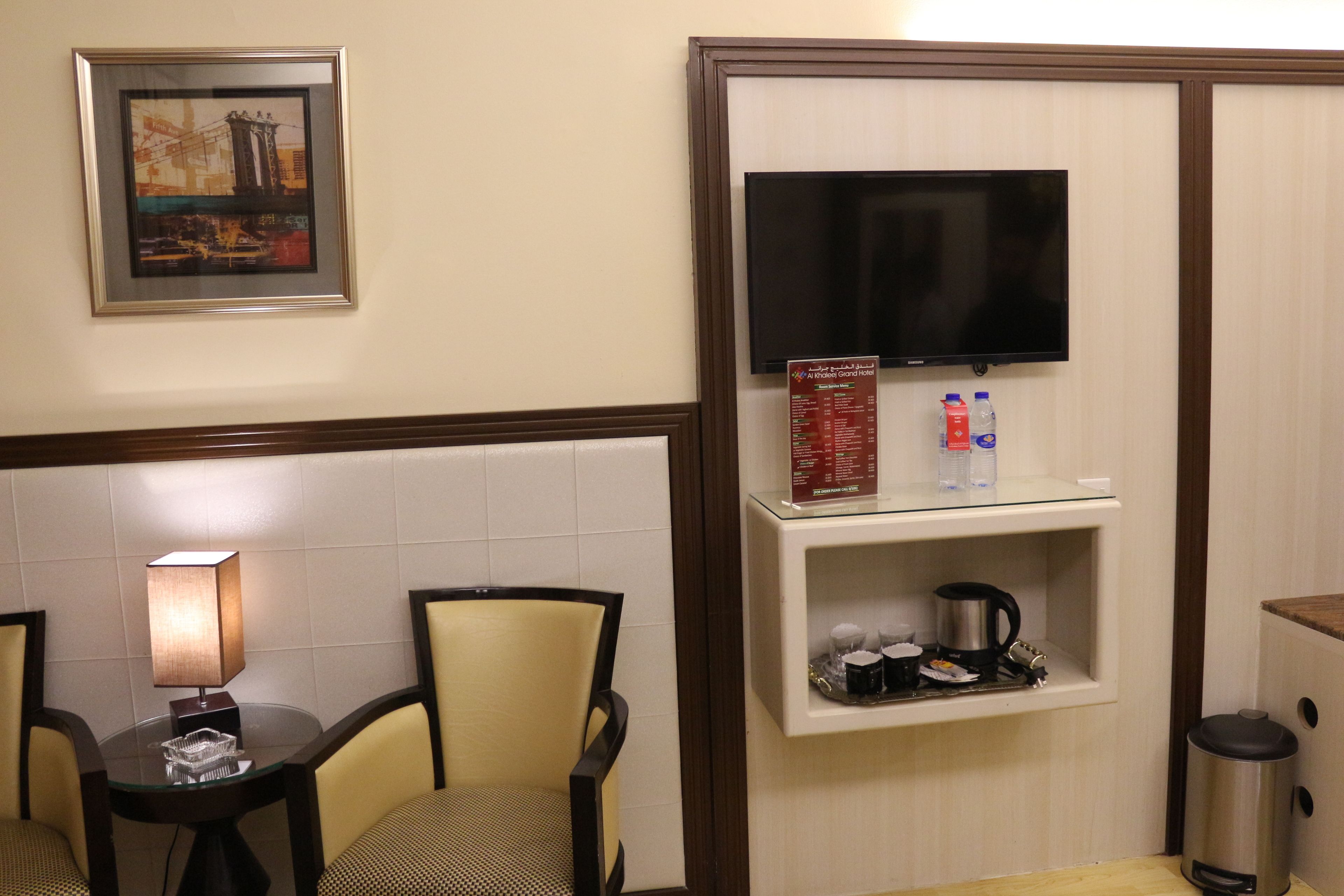 Habitación de hotel con mesa de comedor, sillas, lámpara, televisión, estante con cafetera y cubo de basura.