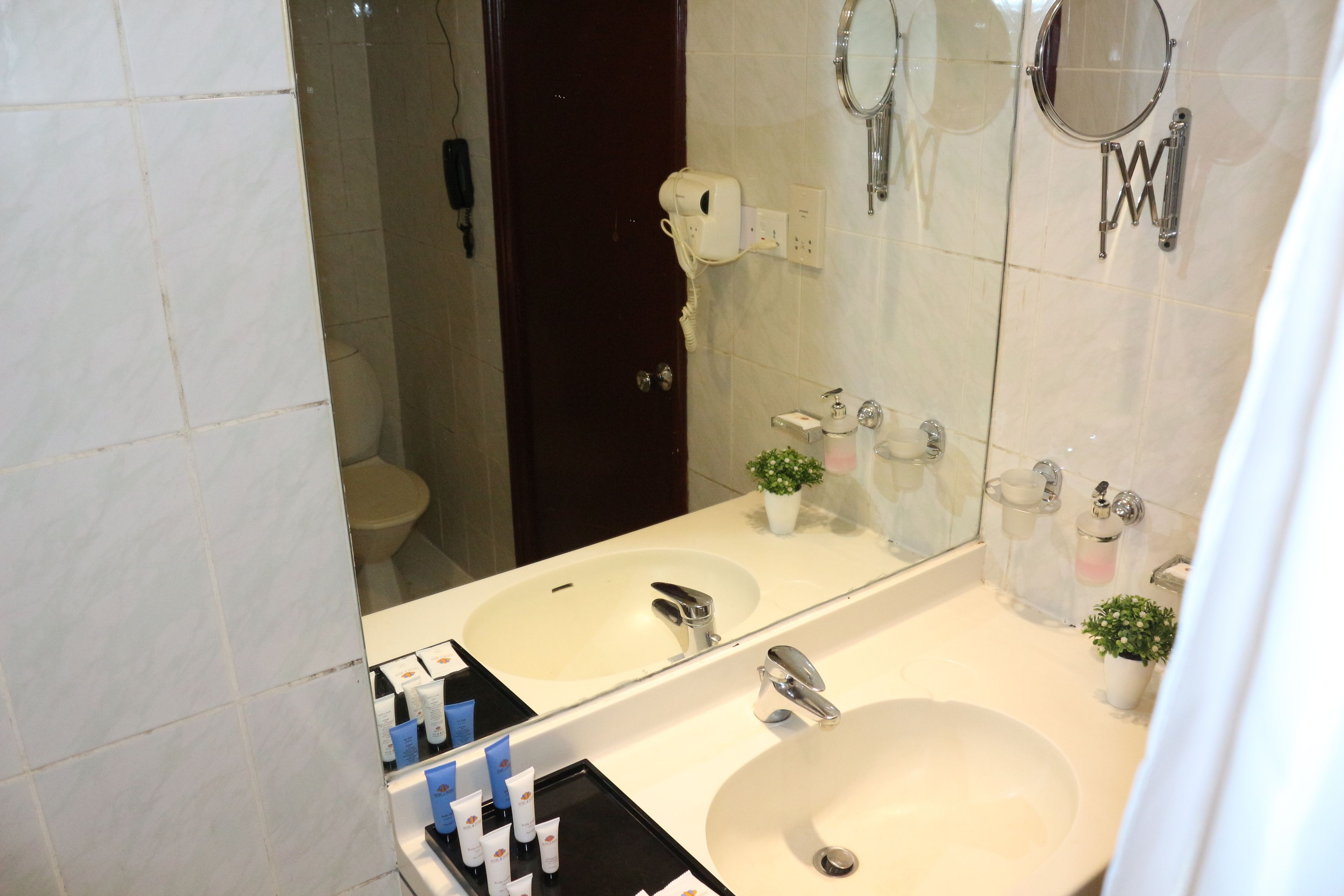 Une salle de bain avec un grand miroir, deux lavabos, des toilettes et un sèche-cheveux fixé au mur.