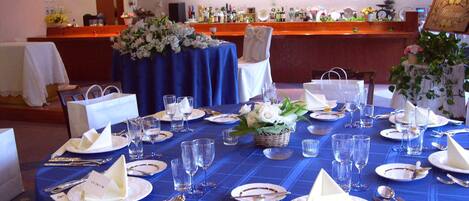 A banquet table set with white plates, silverware, and clear glasses, with a blue tablecloth and a floral centerpiece.