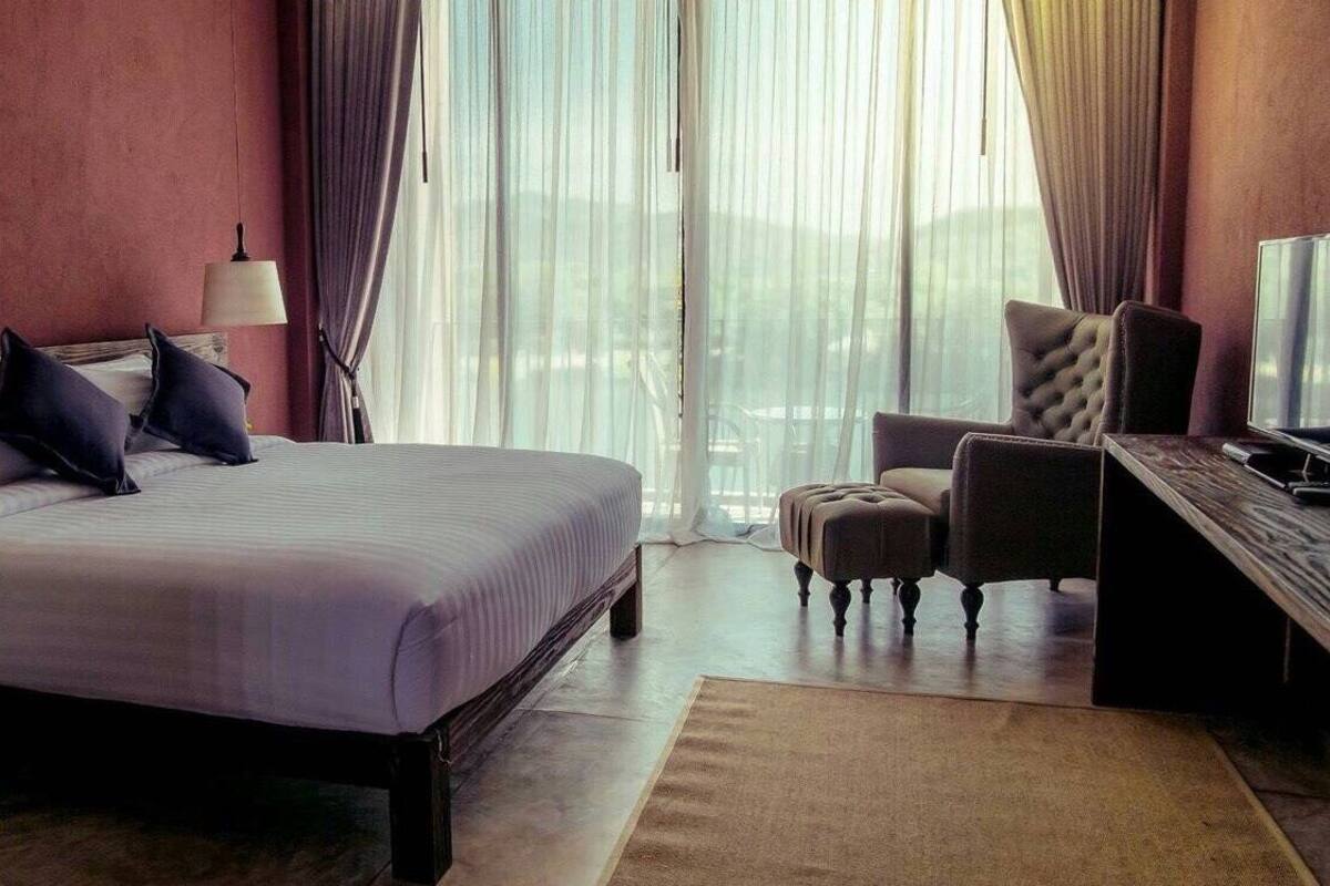 A hotel room with a large bed, a desk with a computer, a chair, and a view of the outdoors through sheer curtains.
