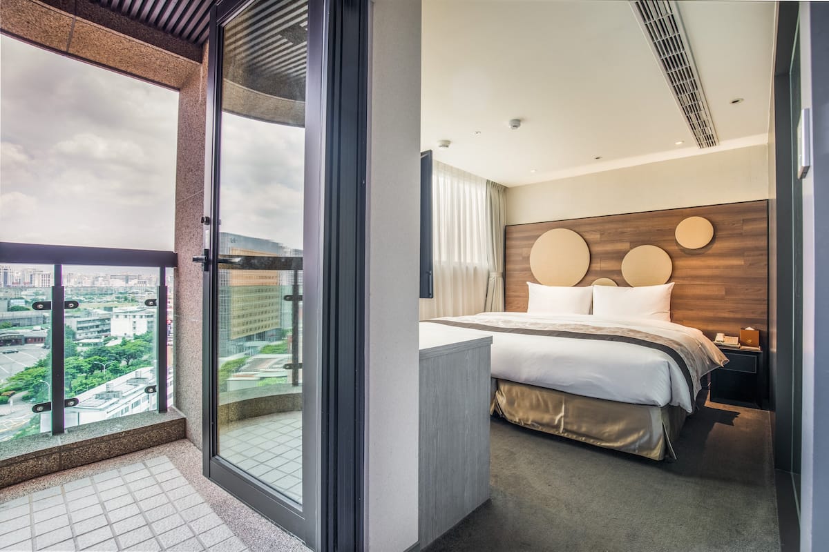 Ein modernes Hotelzimmer mit einem großen Bett, einem hölzernen Kopfteil, einem Nachttisch und Blick auf die Stadt vom Balkon.
