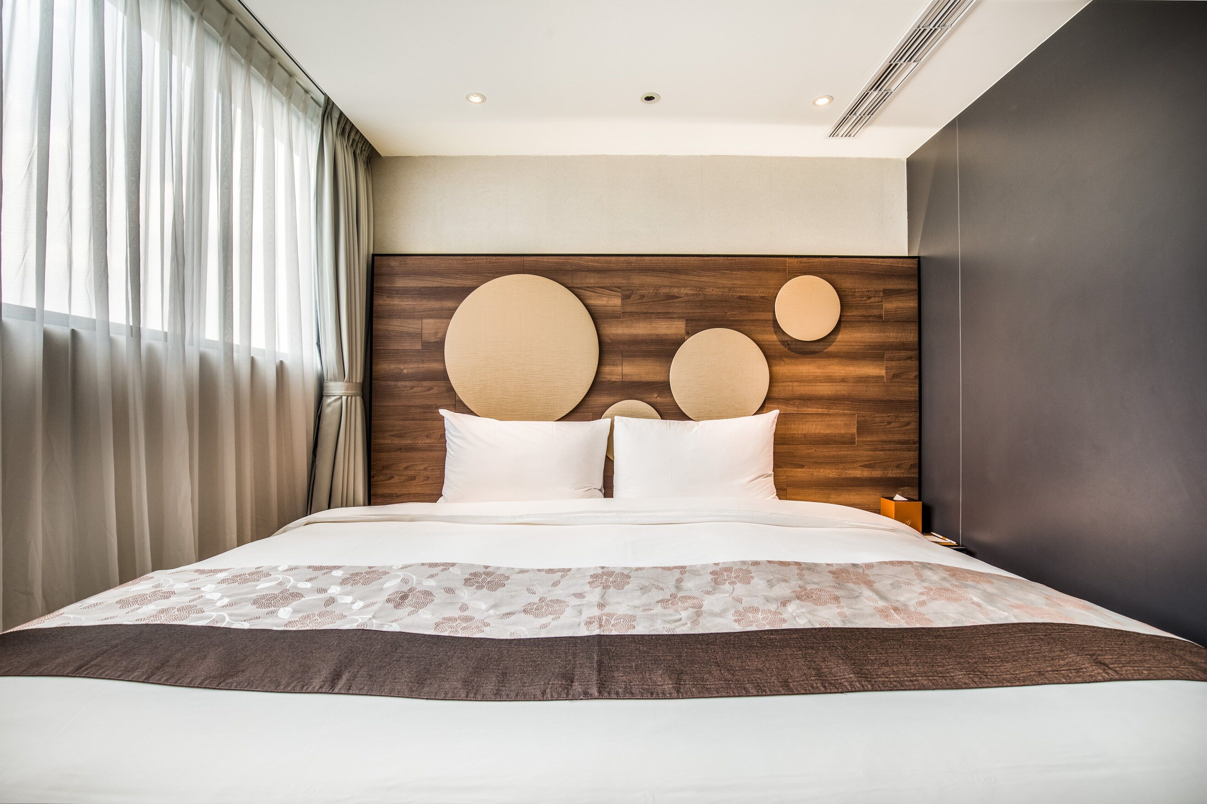 Habitación de hotel moderna con una cama grande, cabecera de madera con detalles circulares y vistas de la ciudad a través de cortinas translúcidas.