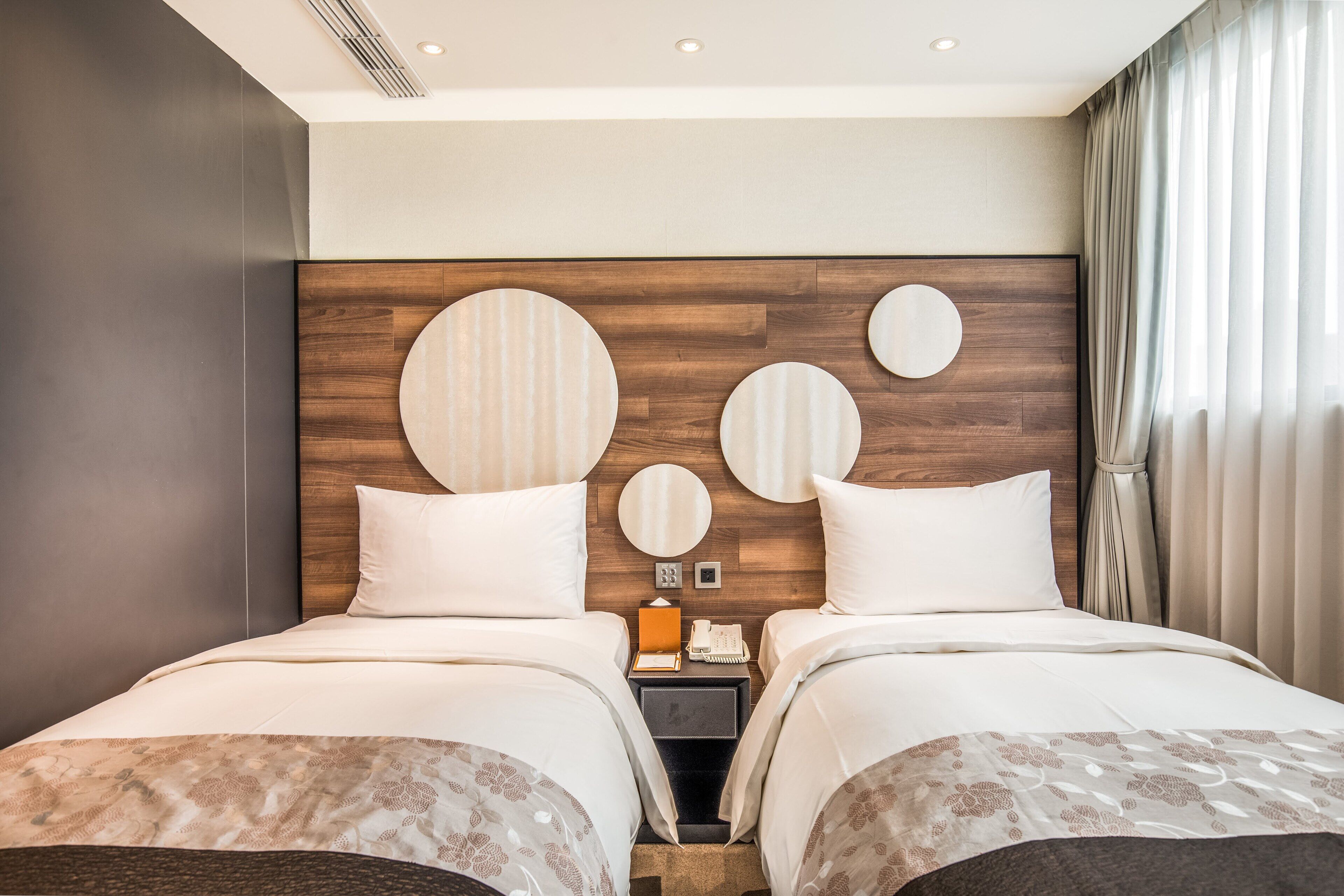A modern hotel room with two beds, a wooden headboard, and a nightstand with a phone and a lamp.