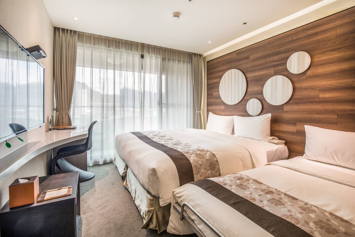 Ein Hotelzimmer mit zwei Betten, einem Schreibtisch und einem großen Fenster mit Vorhängen.