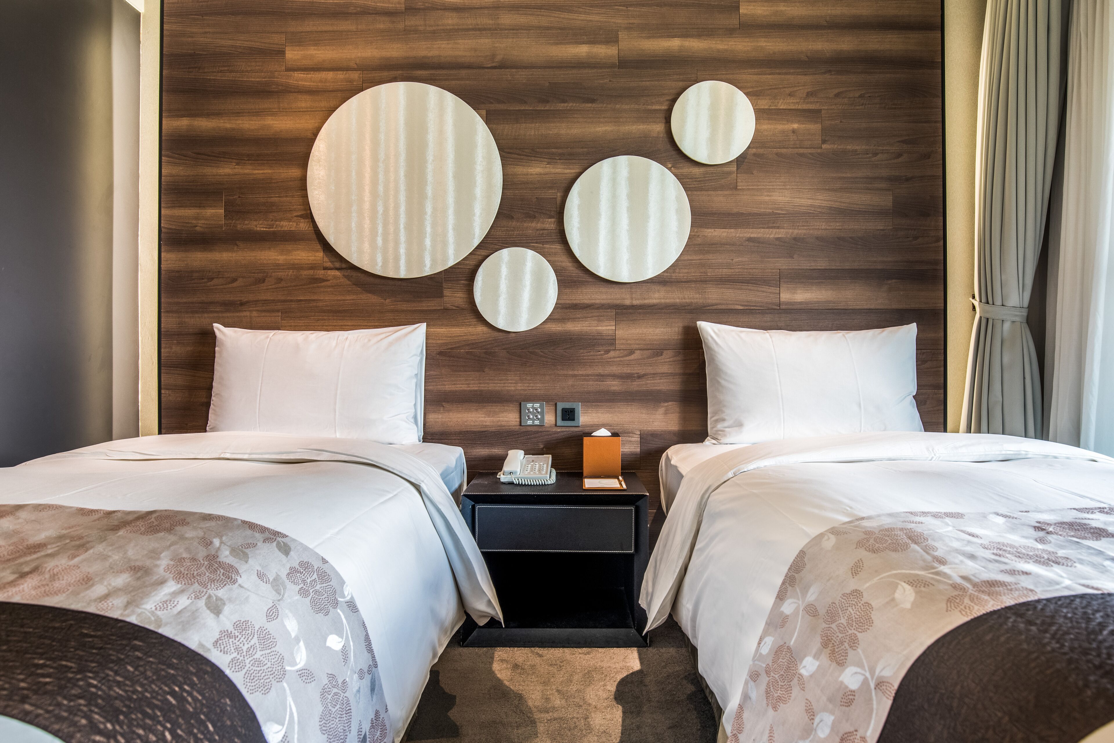 A hotel room with two beds, a wooden headboard, a nightstand with a phone and lamp, and a wall with circular mirrors.