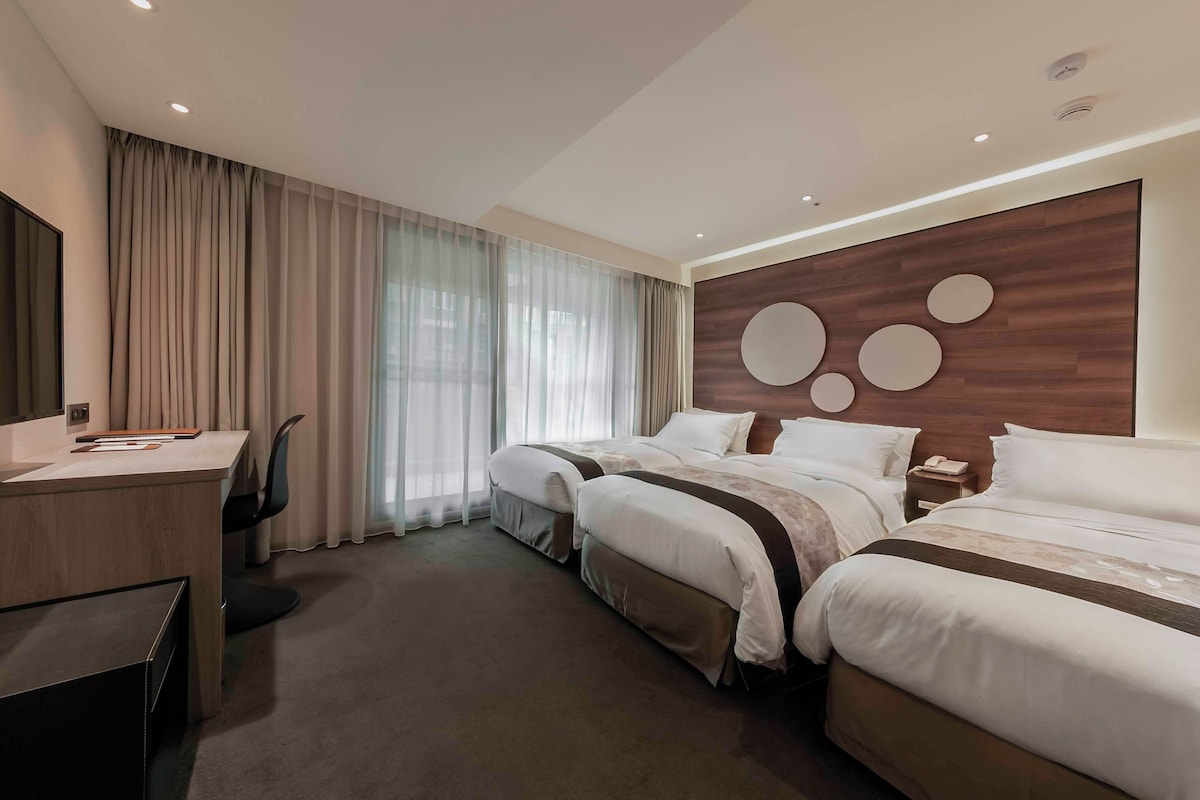 A hotel room with two beds, a desk, a chair, and a large window with curtains.
