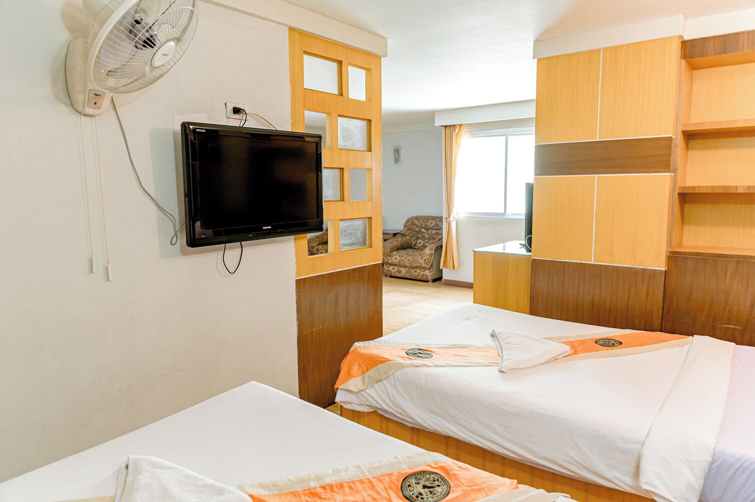 A hotel room with two beds, a TV mounted on the wall, a fan, and wooden furniture.