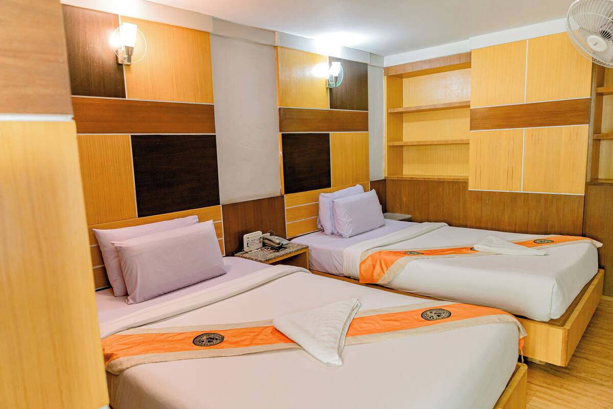 A hotel room with two beds, wooden headboards, a nightstand with a phone, and a wall-mounted fan.