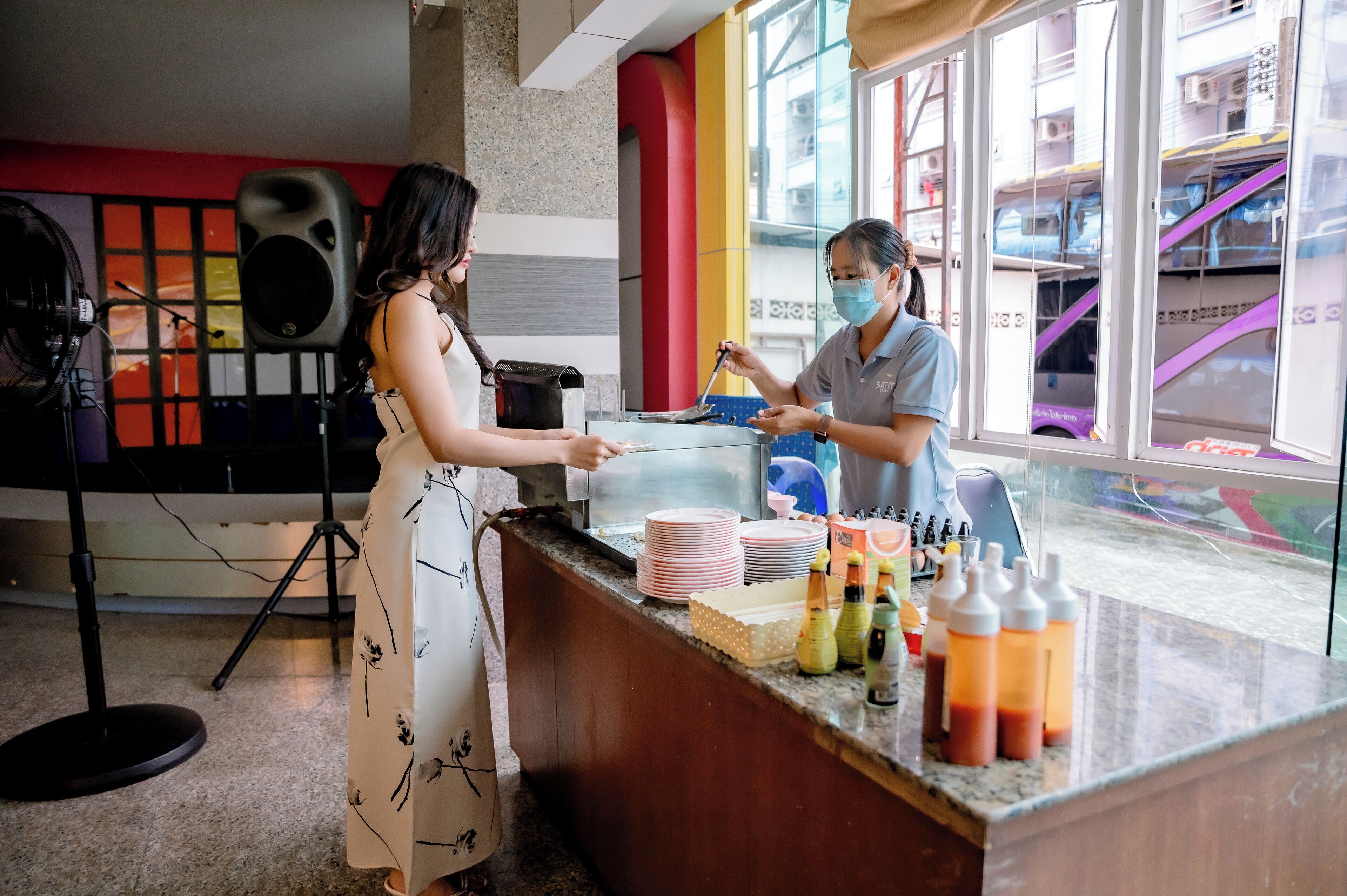 A counter with a woman and a staff member at a restaurant. There are plates, bottles, and a microphone in the background.