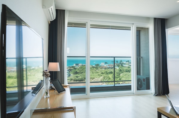 A modern hotel room with a large balcony, a desk with a laptop, and a view of the sea and greenery. - Sally's Jeju (Jeju City)