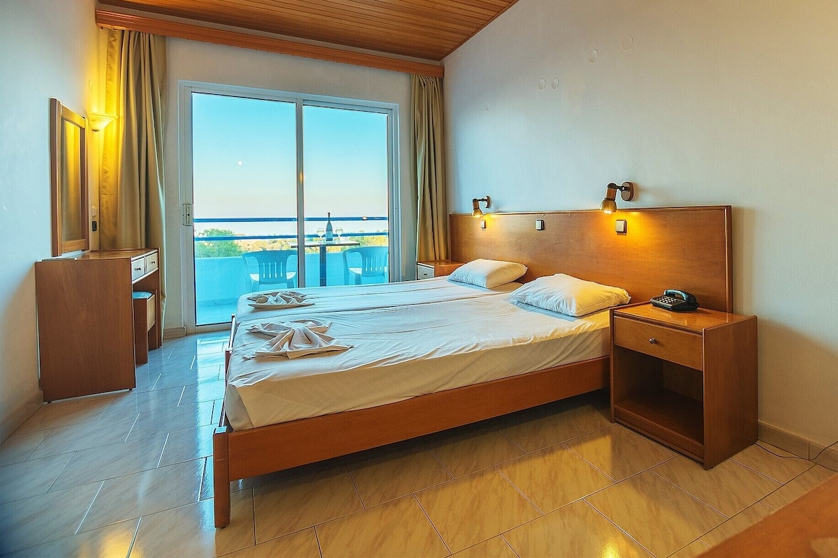 A hotel room with a wooden ceiling, a large bed with white linens, a nightstand with a phone, and a view of the beach through the window.