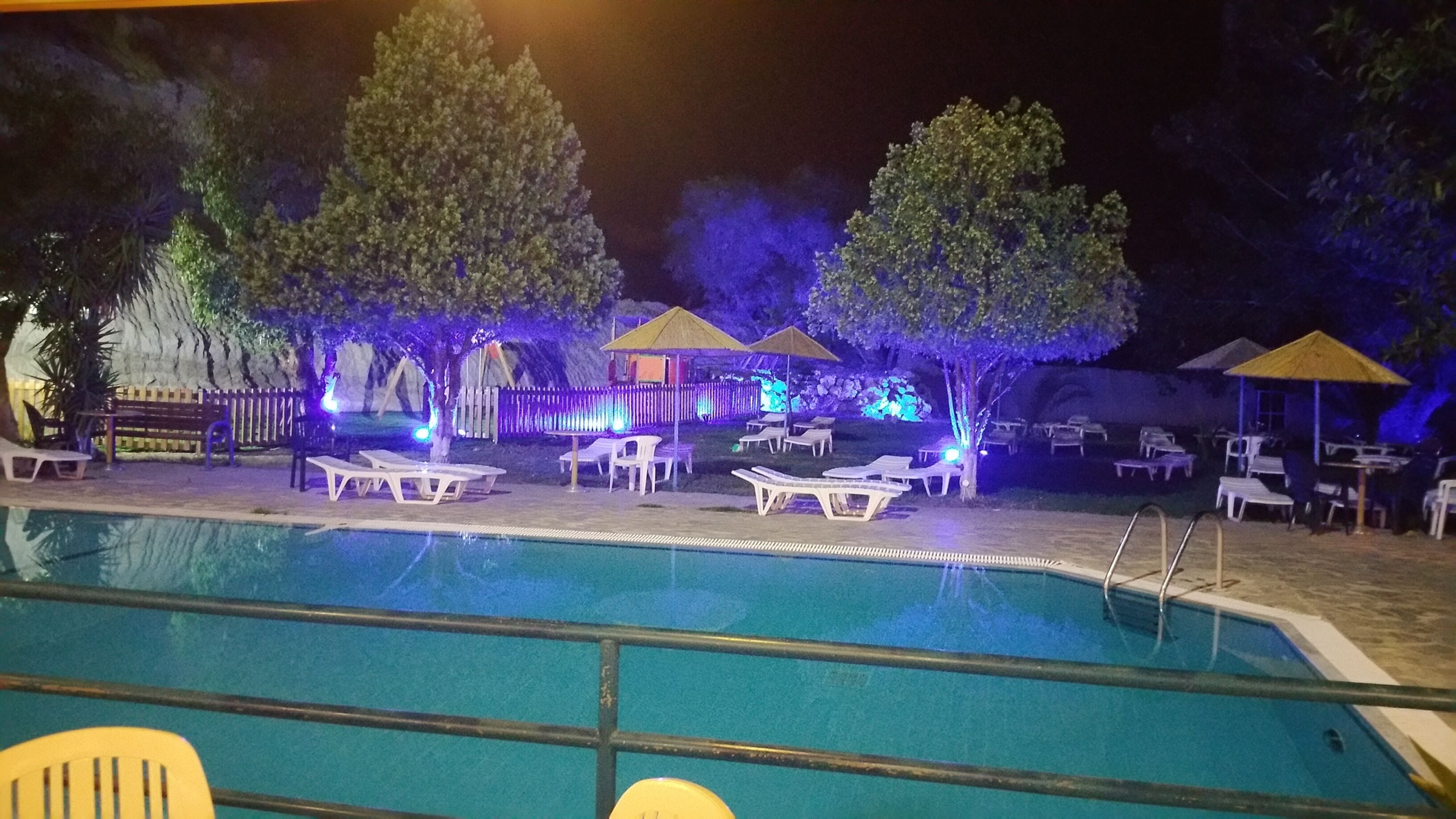 A nighttime pool area with lounge chairs and umbrellas, surrounded by trees and a fence.