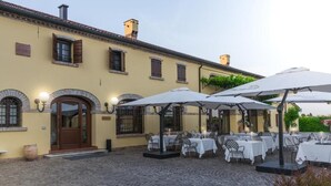 Un espacio exterior para comer con mesas y sombrillas, un patio empedrado y un edificio con fachada amarilla y ventanas de arco.