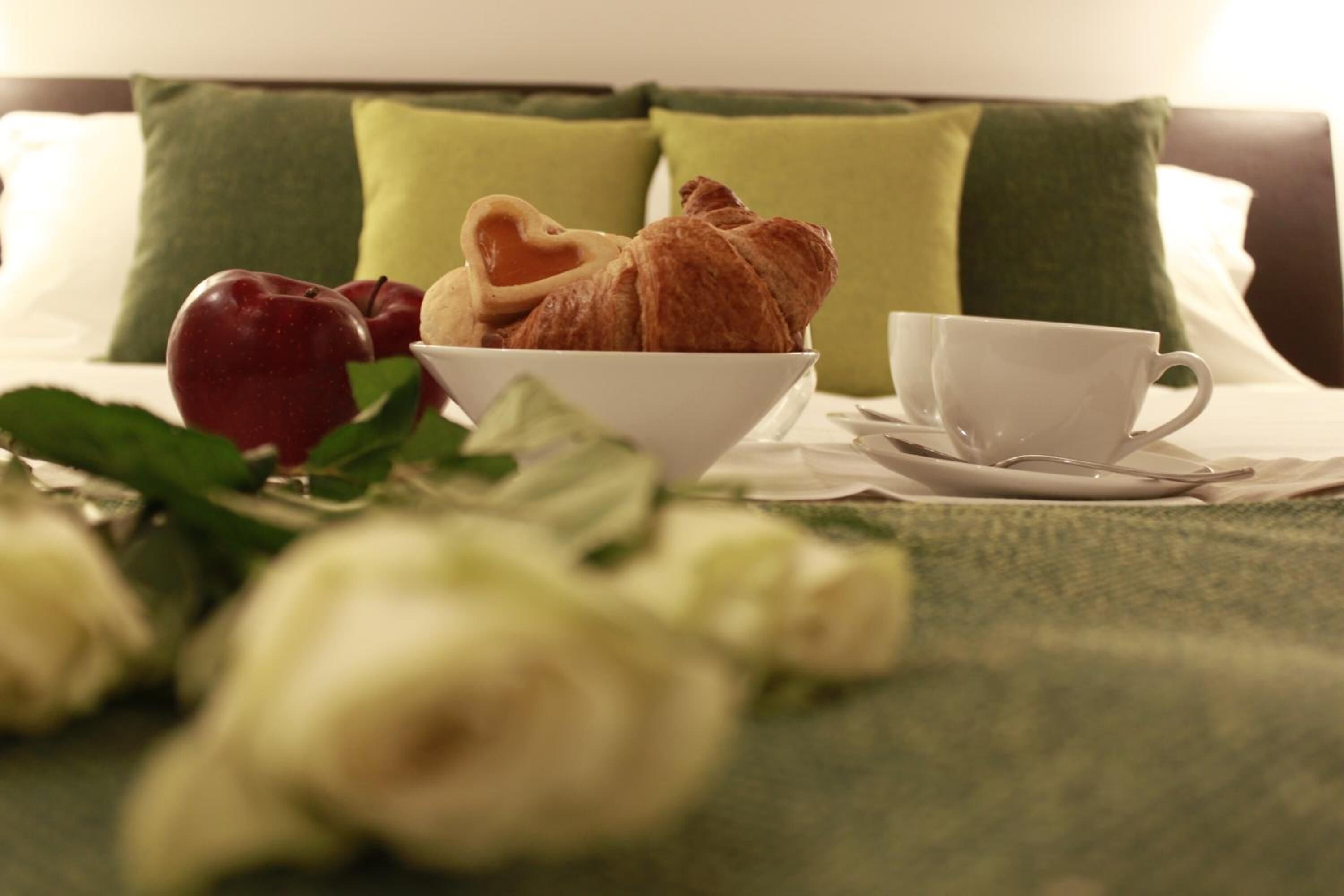 Ein Bett mit einem grünen Kissen, eine Schüssel mit Croissants und Äpfeln sowie eine Tasse auf einem Tisch.