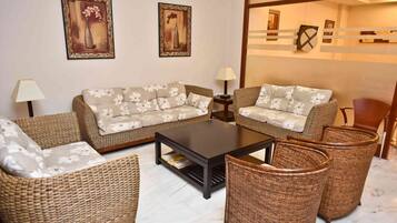 A living room with a sofa set, coffee table, and wicker chairs. There are two wall-mounted lamps and two framed artworks.