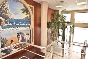 Vestíbulo de hotel con un mural de una escena de playa, una escalera y plantas de interior.