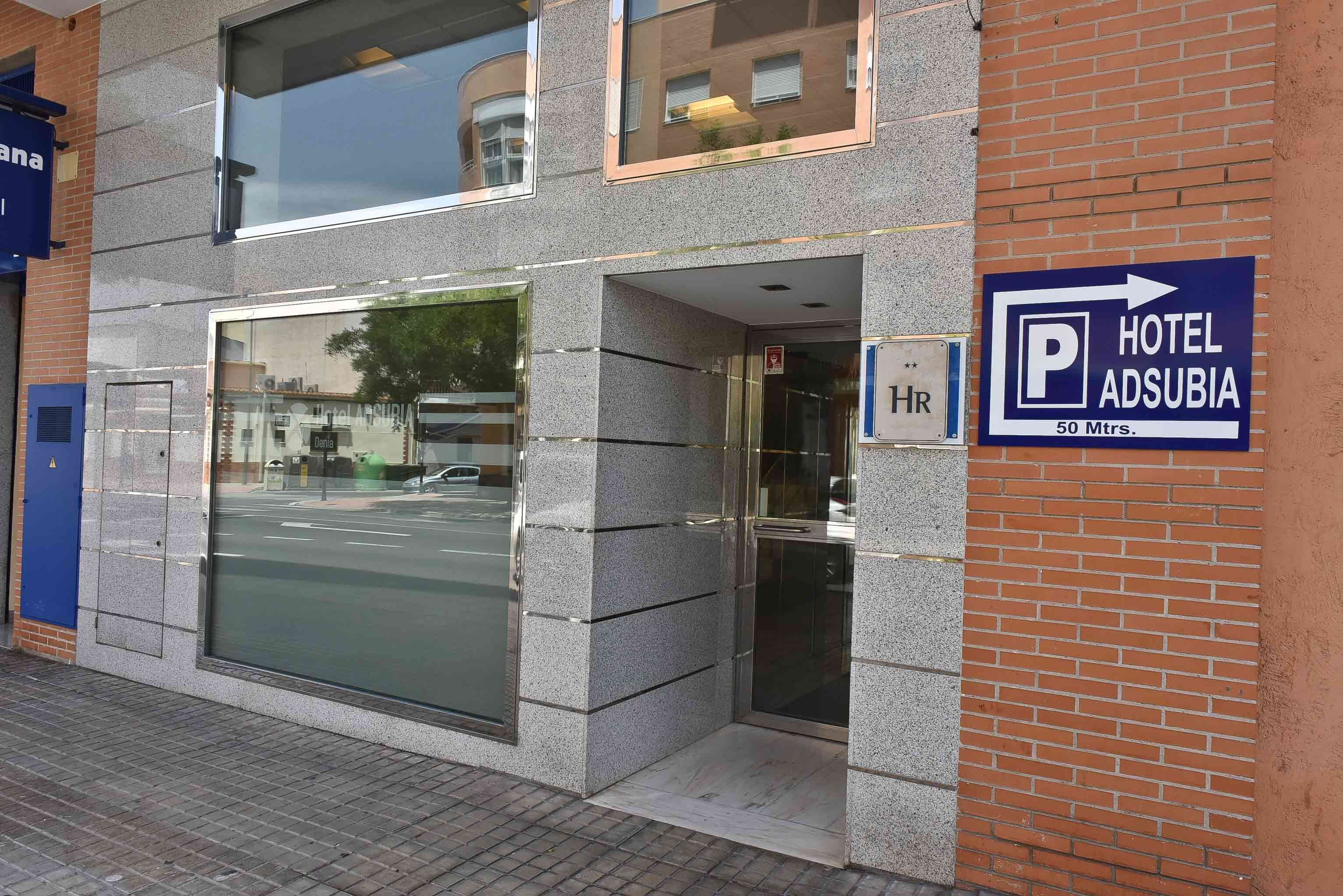 Entrada de un hotel con un amplio ventanal de vidrio, un cartel que indica 'HOTEL ADSUBIA 50 Mtrs.' y una señal de estacionamiento con una flecha.