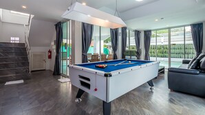 A modern pool table in a well-lit room with a staircase, a sofa, and a view of greenery outside. - Pattaya Pool Villa (Pattaya)