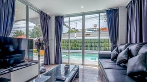 A modern living room with a glass coffee table, a leather sofa, and a TV. Large windows offer a view of a swimming pool and a building in the background. - Pattaya Pool Villa (Pattaya)
