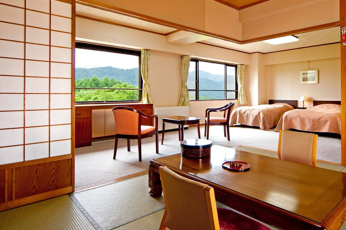 A traditional Japanese room with tatami flooring, a low table, two chairs, a bed with a wooden headboard, and a view of green mountains through large windows.