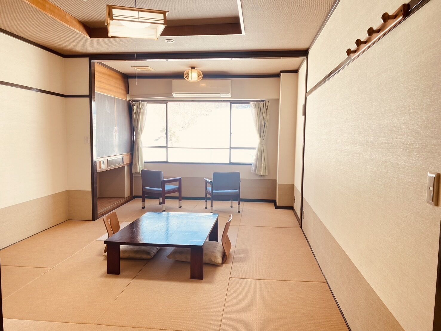 A traditional Japanese room with tatami flooring, a low table, two chairs, a sliding door, and a window with curtains.