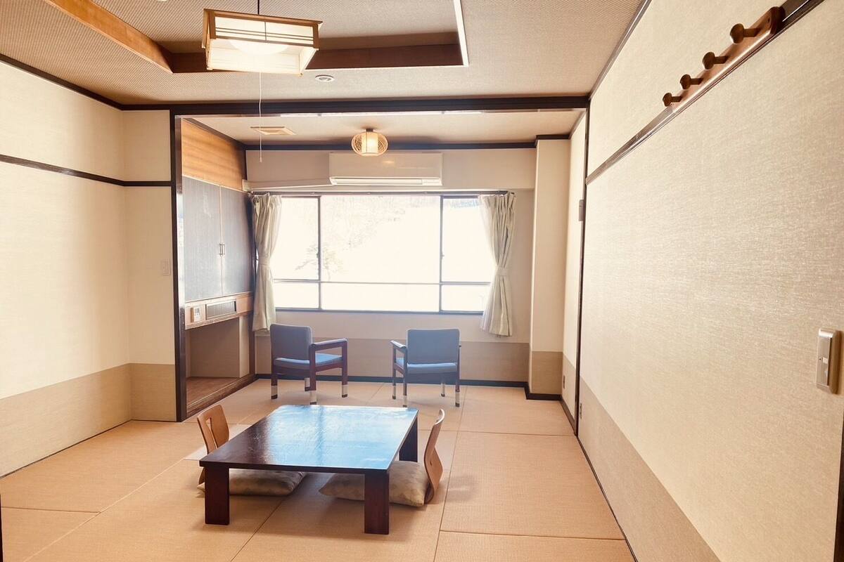 A traditional Japanese room with tatami flooring, a low table, two chairs, a sliding door, and a window with curtains.