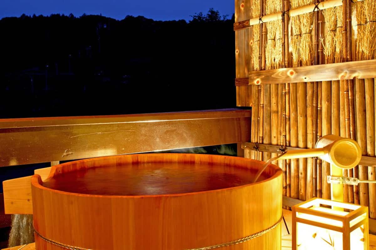 A wooden hot tub with a wooden bench and a small stool, set against a bamboo wall.