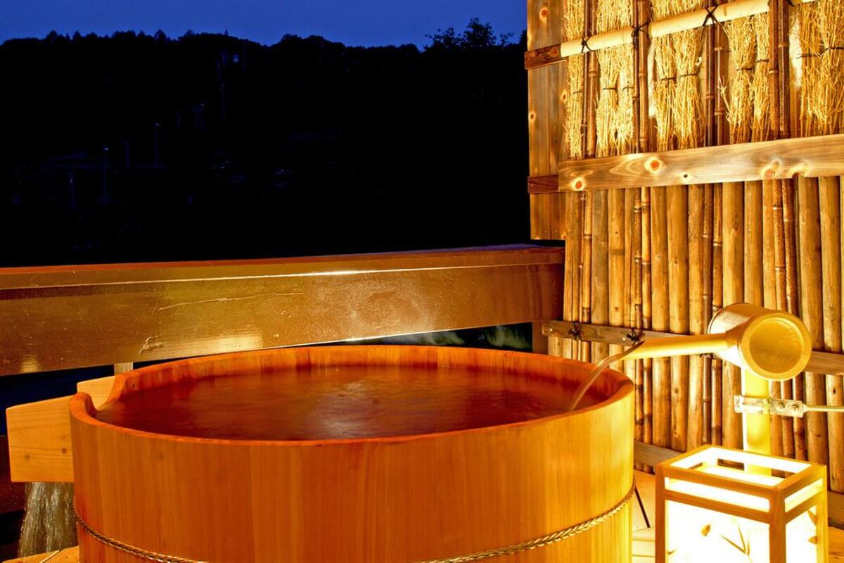 A wooden hot tub with a wooden bench and a small stool, set against a bamboo wall.