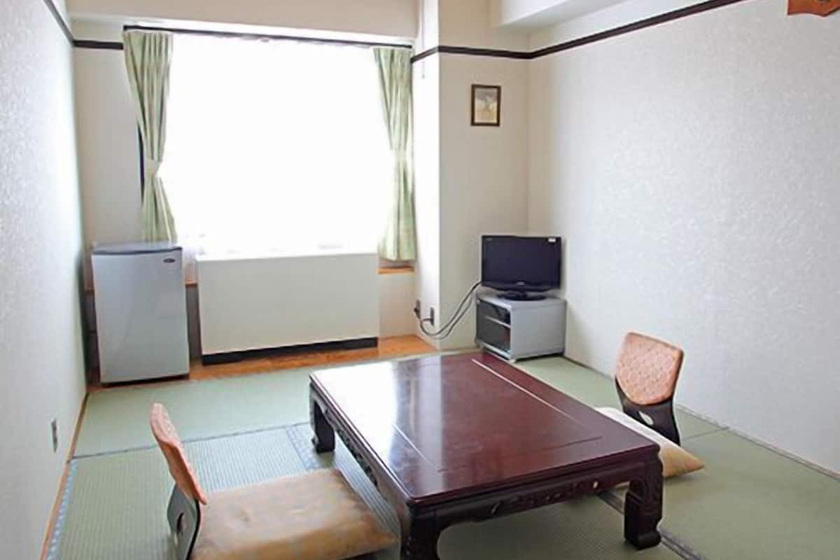 A room with tatami flooring, a wooden table, a television, a refrigerator, and a window with curtains.