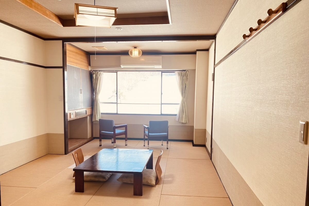 A traditional Japanese room with tatami flooring, a low table, two chairs, a sliding door, and a window with curtains.