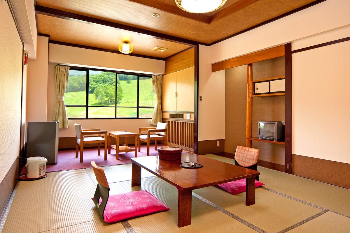 A traditional Japanese room with tatami flooring, a low table, chairs, a television, and a view of greenery outside.
