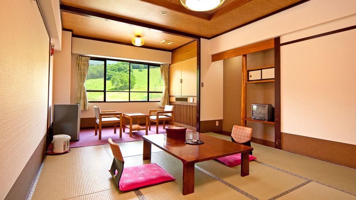 A traditional Japanese room with tatami flooring, a low table, chairs, a television, and a view of greenery outside.