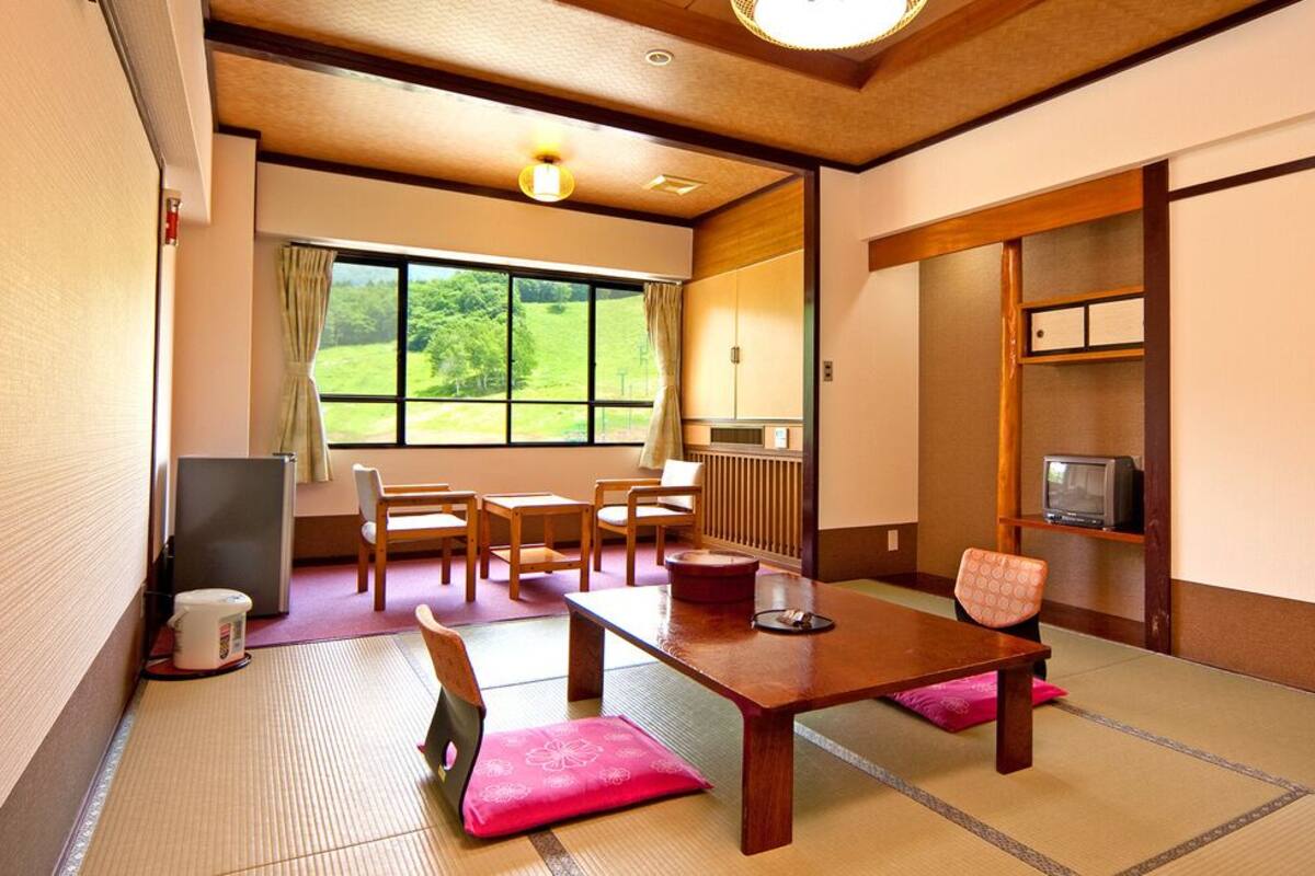 A traditional Japanese room with tatami flooring, a low table, chairs, a television, and a view of greenery outside.