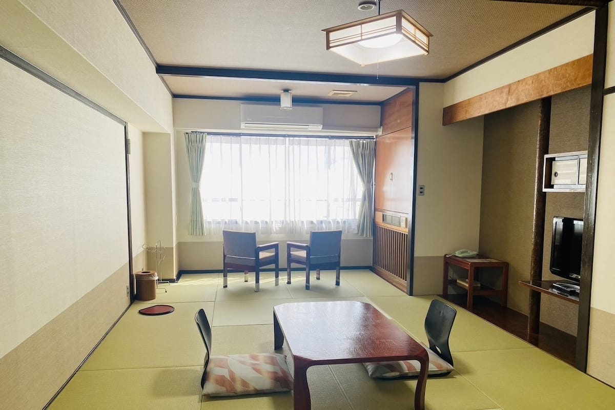 A traditional Japanese-style room with tatami flooring, a low table, chairs, a television, and a sliding door.