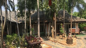 A courtyard with a tiled floor, palm trees, and a building with a corrugated metal roof. - Sam's House Guesthouse (Kanchanaburi)