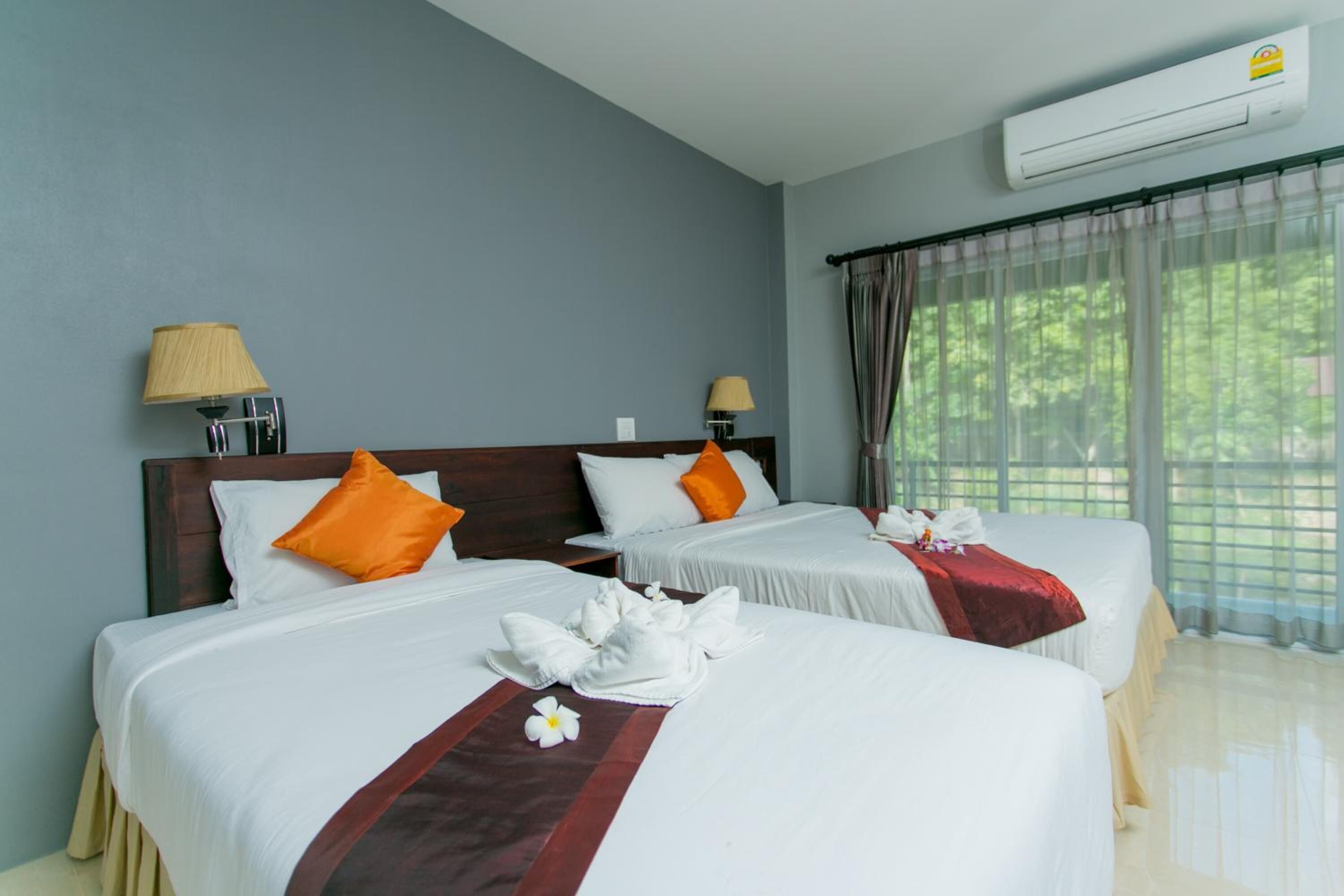 Habitación de hotel con dos camas, cada una con ropa blanca y almohadas anaranjadas, cabecero de madera, pared color beige y un equipo de aire acondicionado.