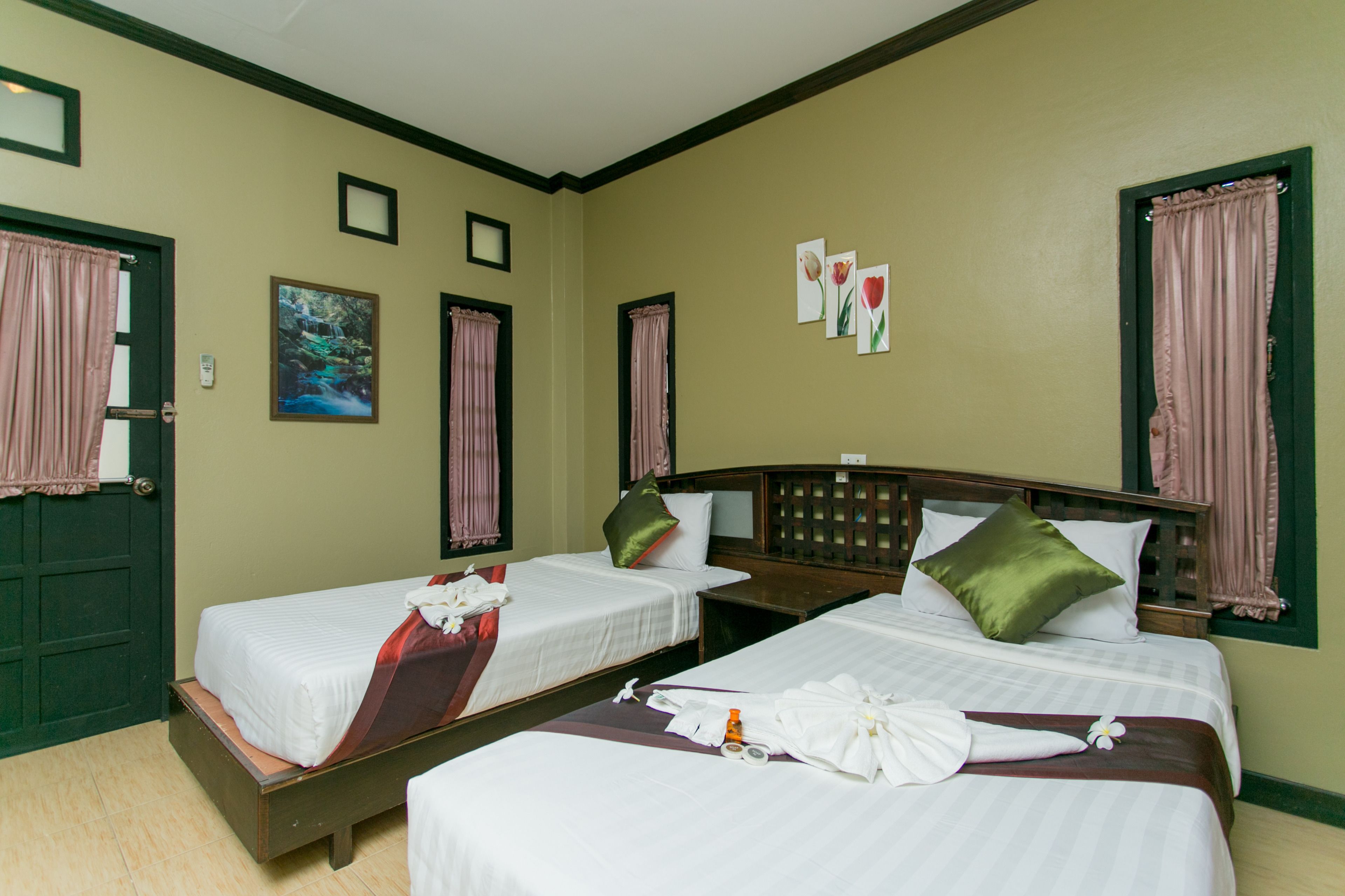 A hotel room with two single beds, each with a pillow and a blanket. There are three framed pictures on the wall, and a green door with a window.