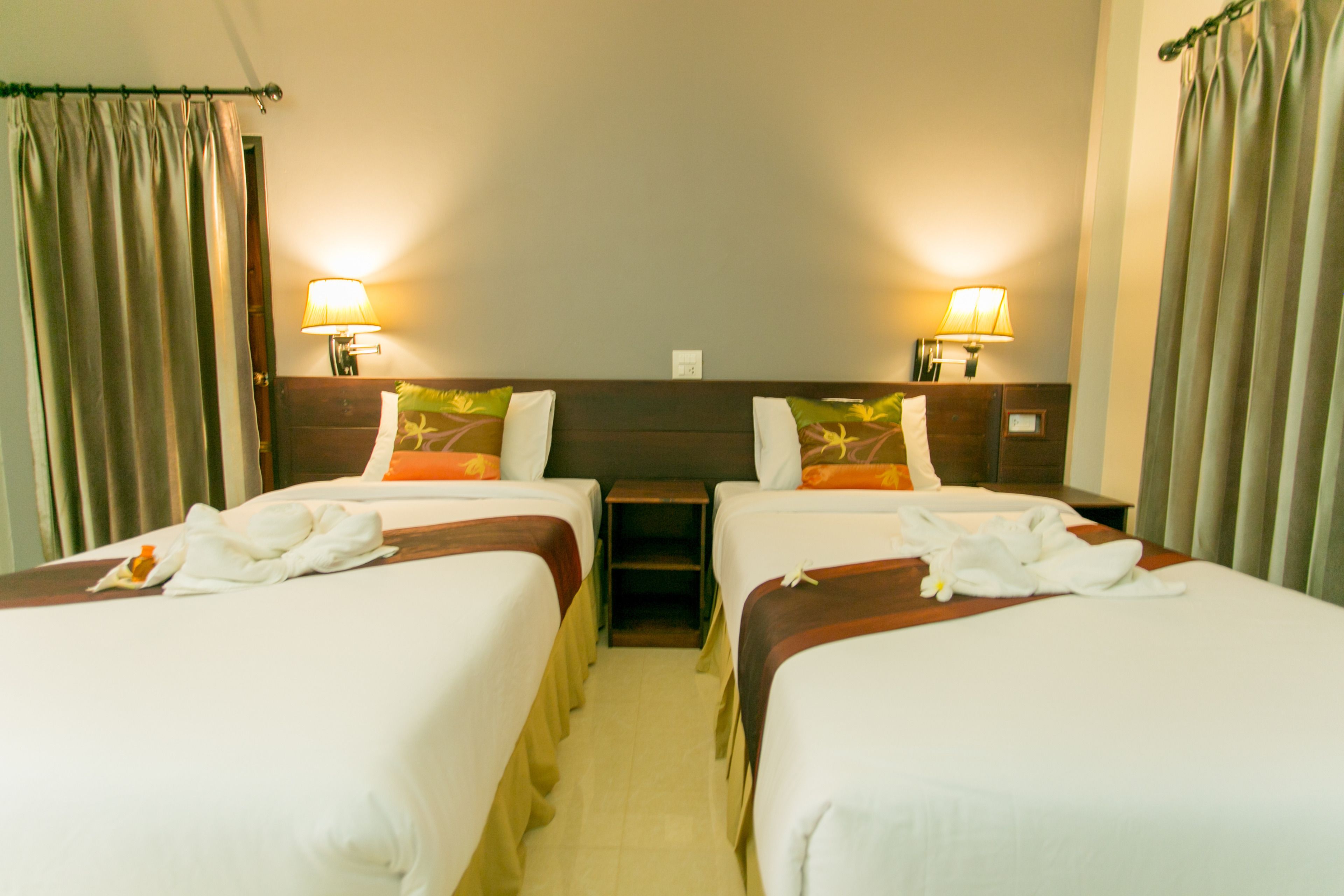 Habitación de hotel con dos camas, cada una con una almohada y una toalla. Hay dos lámparas de noche y una mesita entre las camas.