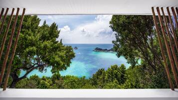 Blick durch ein Fenster mit Bambusrollos auf ein türkisblaues Meer, üppiges Grün und einen klaren Himmel.