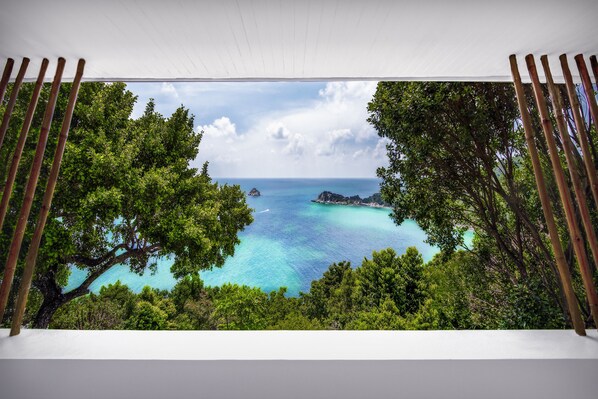Une vue à travers une fenêtre ornée de stores en bambou, donnant sur une mer turquoise, une végétation luxuriante et un ciel dégagé.