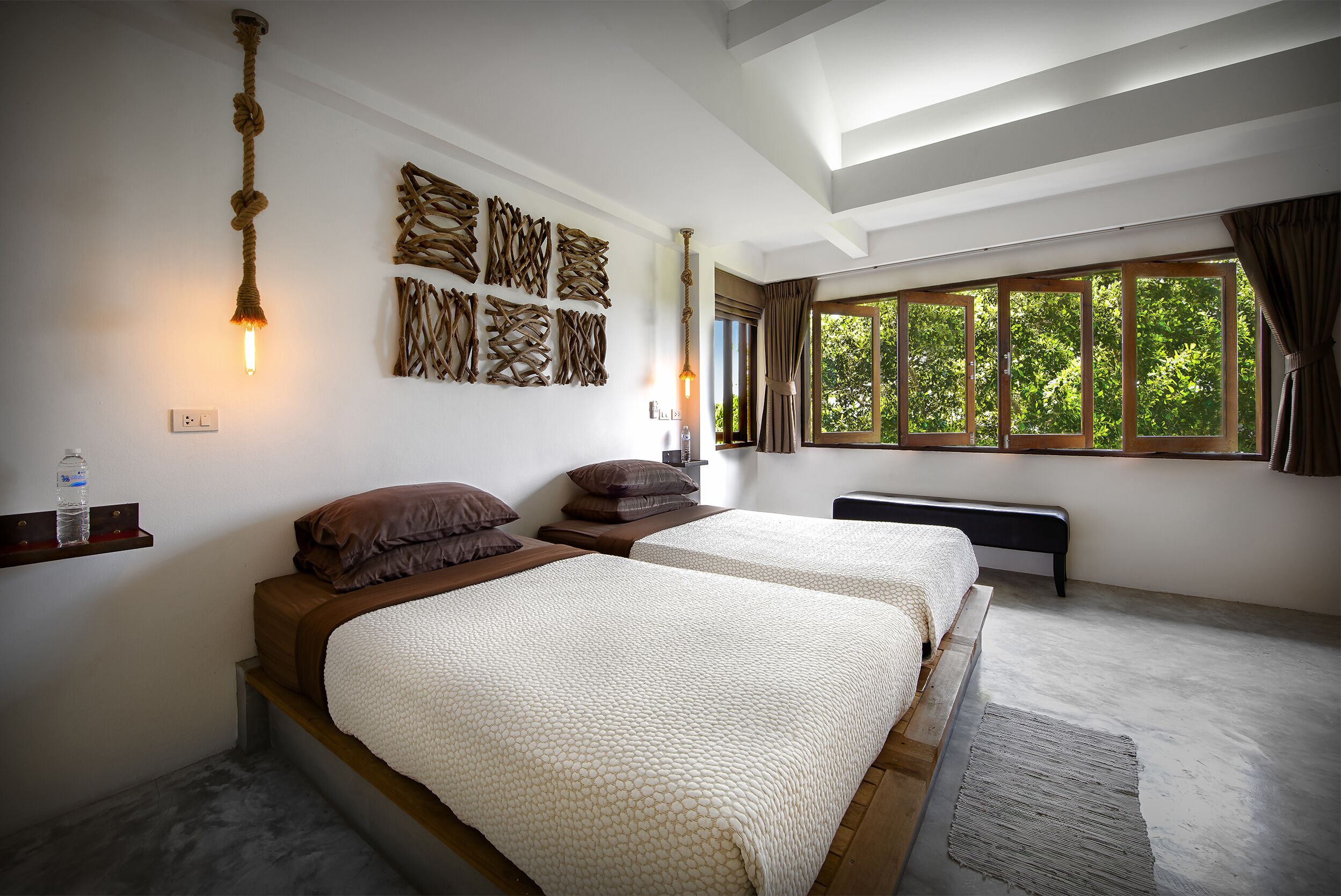 A bedroom with two beds, a wooden bench, a wall-mounted light fixture, and a window with curtains.