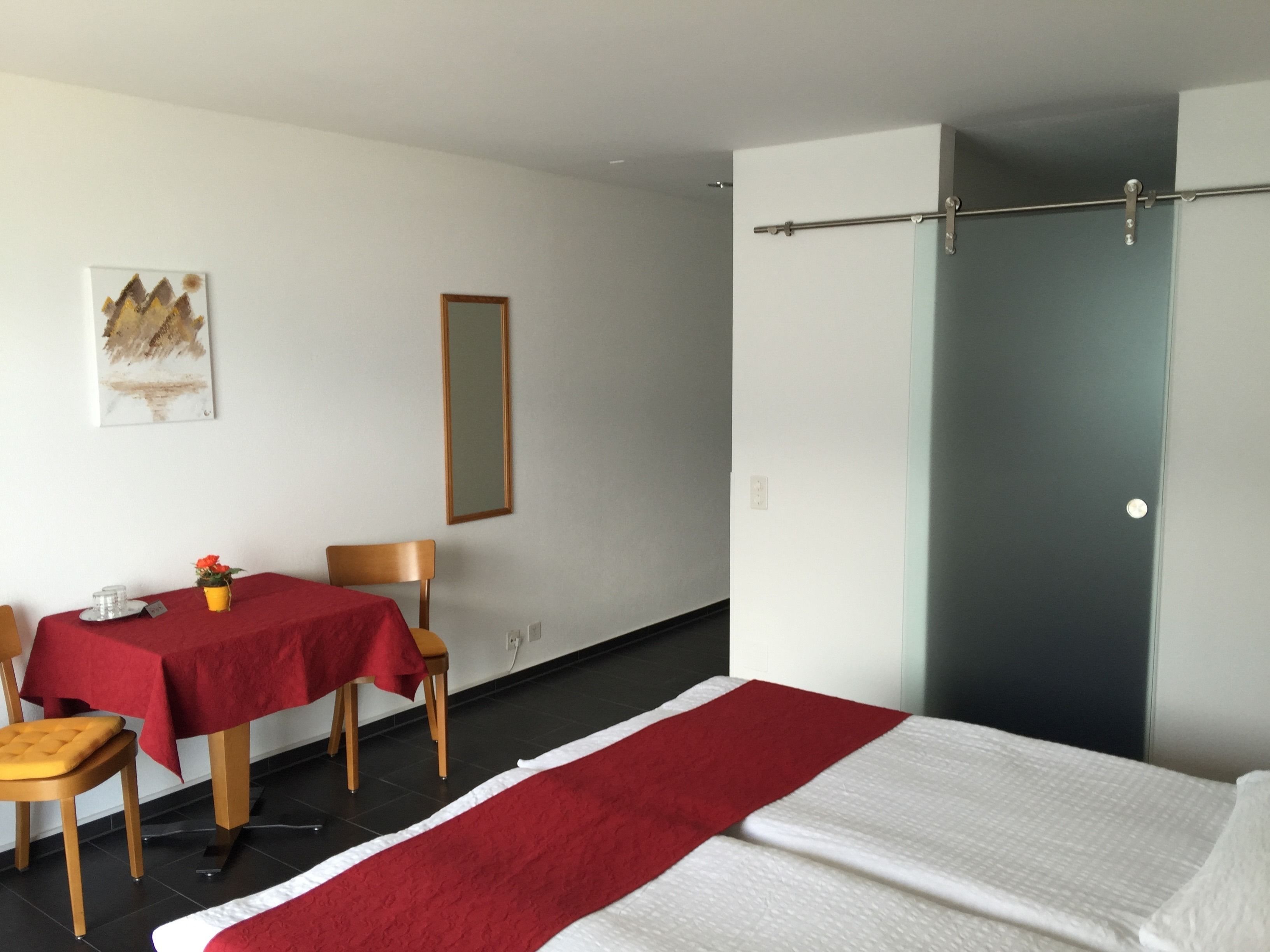 A hotel room with a bed, a table with a red tablecloth, two chairs, a small table with a vase of flowers, a painting on the wall, and a door with a towel rack.