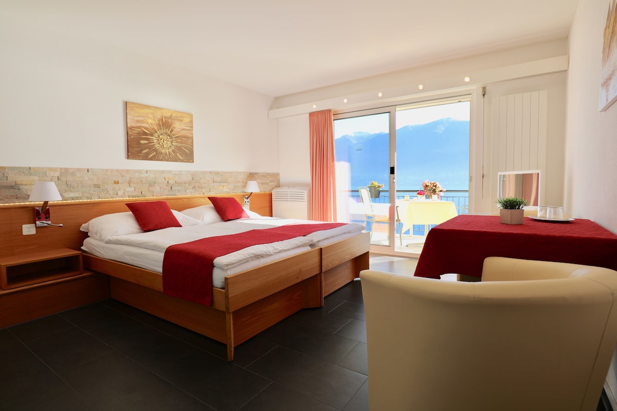 Una habitación de hotel moderna con una cama de madera, una manta roja, una mesita con una planta y vistas a las montañas desde las puertas del balcón.