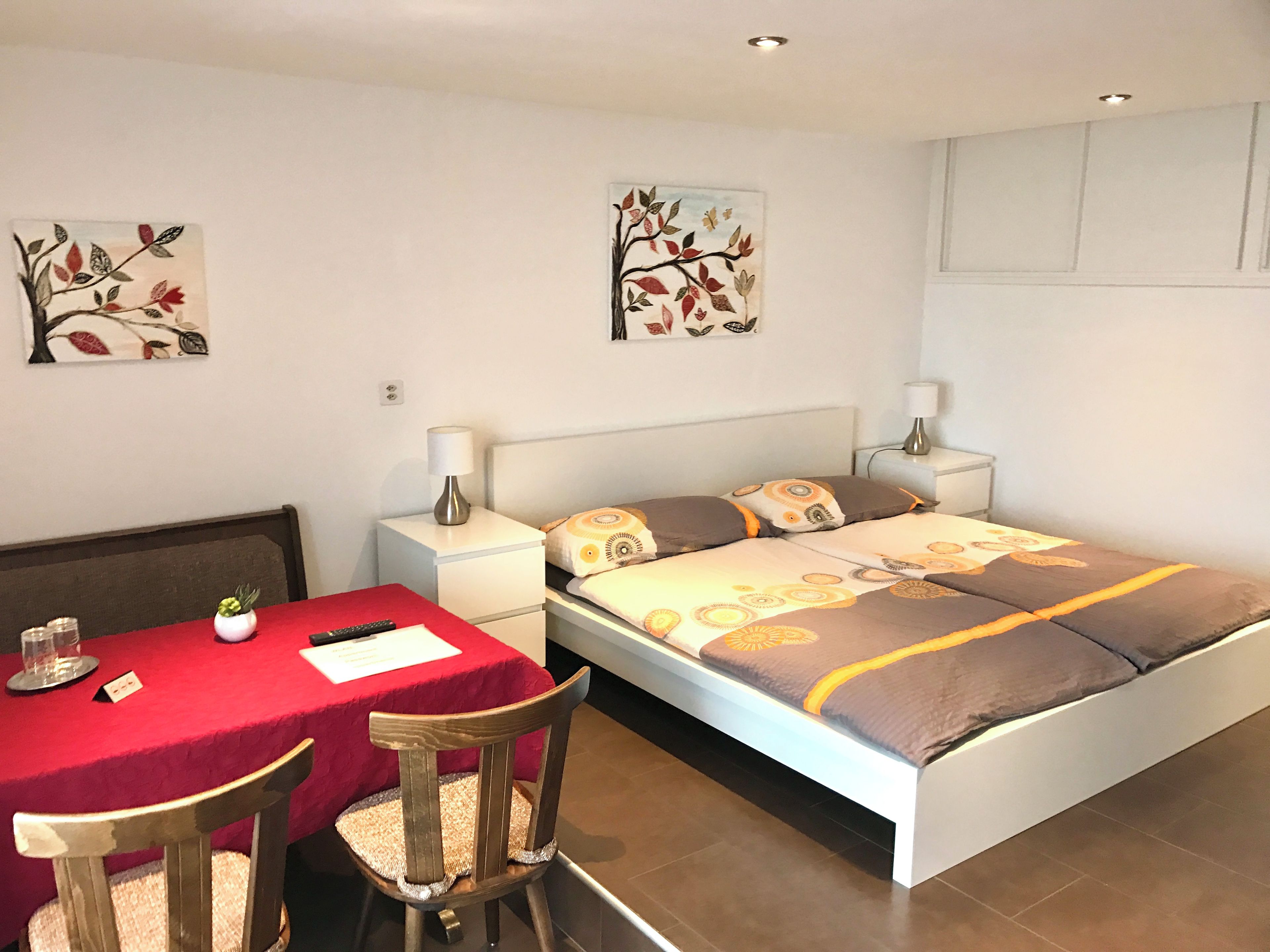 A hotel room with a bed, bedside tables, a desk with chairs, and artwork on the walls.