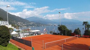 Una cancha de tenis con red, rodeada de vegetación y vistas a un lago y montañas.
