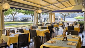 Ein Restaurant im Freien mit Tischen unter gelben Sonnenschirmen, Korbstühlen und Blick auf geparkte Fahrzeuge und Bäume.