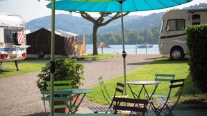 Ein Campingplatz mit Tisch und Stühlen unter einem großen Sonnenschirm, ein Wohnmobil und ein Boot im Hintergrund auf dem Wasser.
