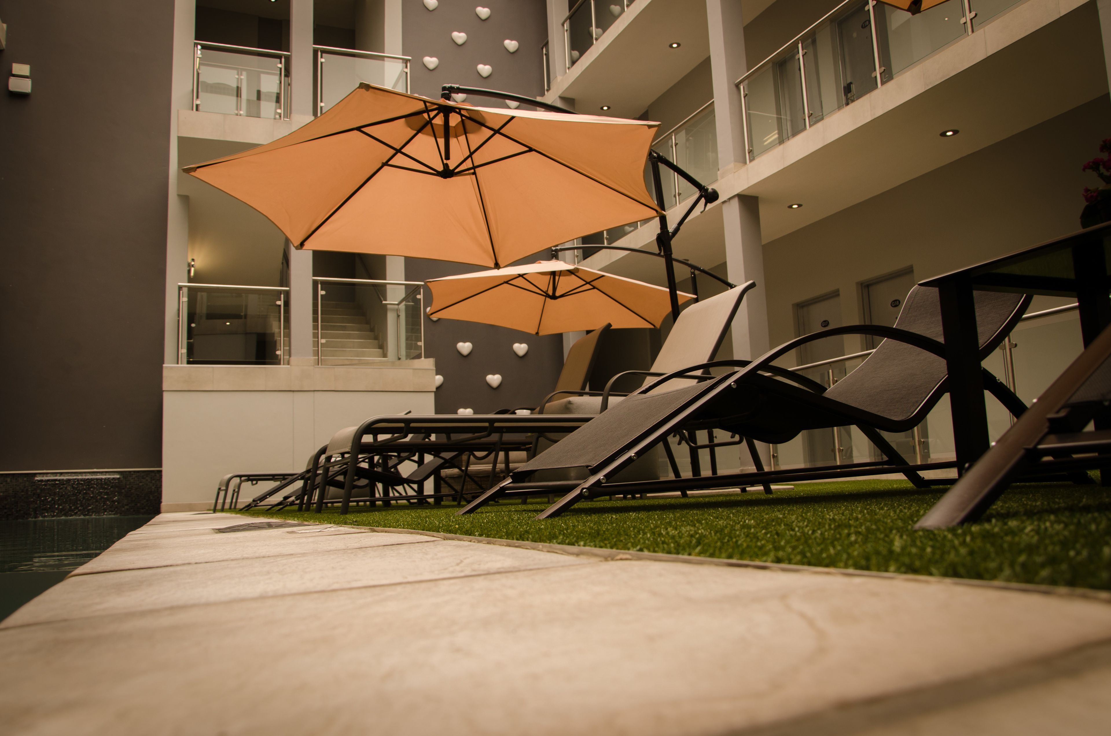 A modern outdoor seating area with two lounge chairs and two large umbrellas, a pool, and a building with multiple floors and heart-shaped decorations.
