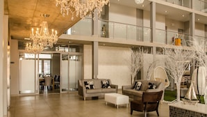 Een ruime lobby met een hoog plafond, een kroonluchter, twee banken met kussens, een bijzettafeltje en een decoratieve plantenarrangement.