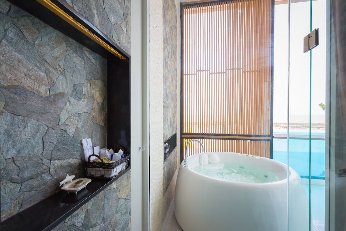 Un baño con pared de piedra, una bañera y ventana con vistas al mar.
